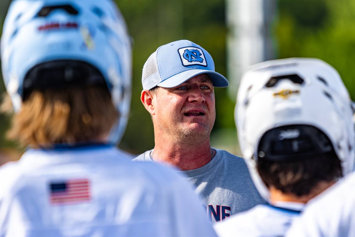 A Team 91 Carolina coach talking to two helmeted players between reps, the players large and out of focus in the foreground.