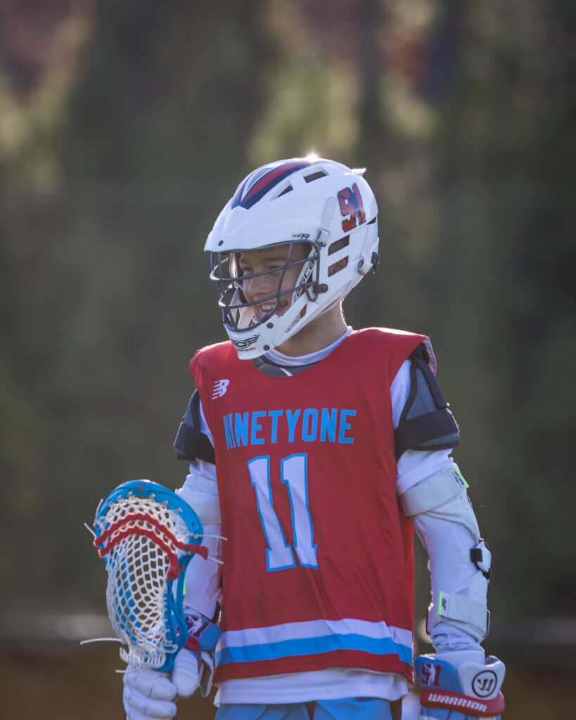 A young Team 91 Carolina player in a red NINETY ONE pinnie and white helmet, smiling on the field with his stick.