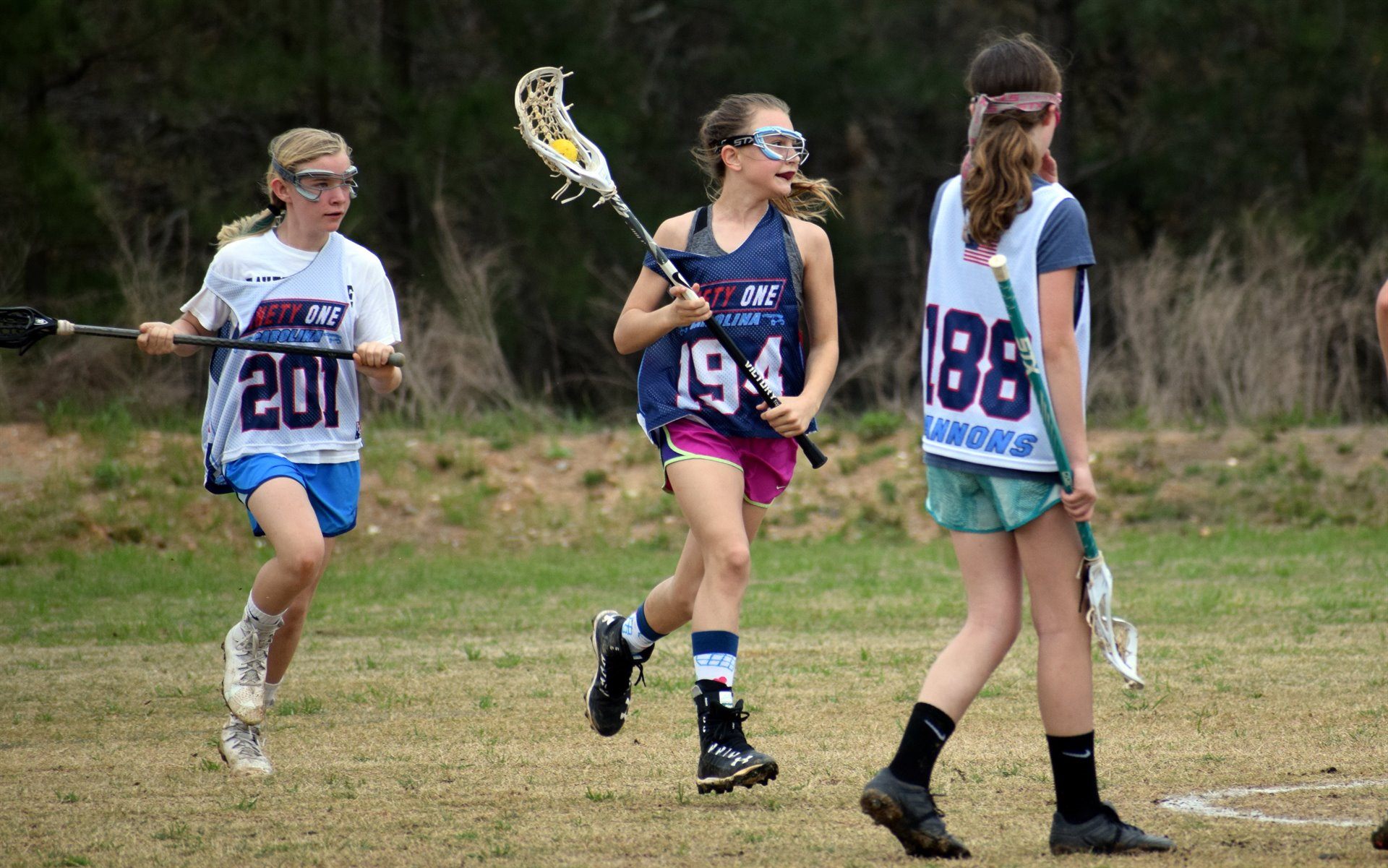 Three young Team 91 Carolina girls players in Ninety One Carolina pinnies moving down a grass field, the player in the middle carrying the ball in her stick.